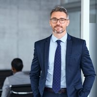 Portrait of handsome mid adult business man standing in modern office. Successful mature entrepreneur in formal clothing looking at camera with satisfaction. Confident man in suit with eyeglasses and beard standing with hands in pocket and looking at camera in office.